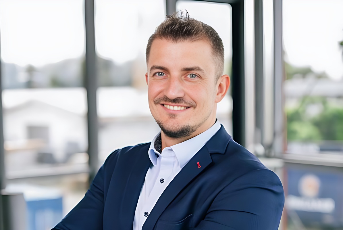 Ein Mann mit kurzem Haar und Bart trägt einen blauen Anzug und ein weißes Hemd, steht vor einem Fenster mit unscharfem Hintergrund und lächelt in die Kamera.